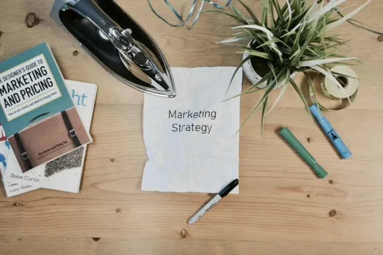 木製の机の上に置かれた「Marketing Strategy」と書かれた紙、マーケティング関連の書籍と観葉植物