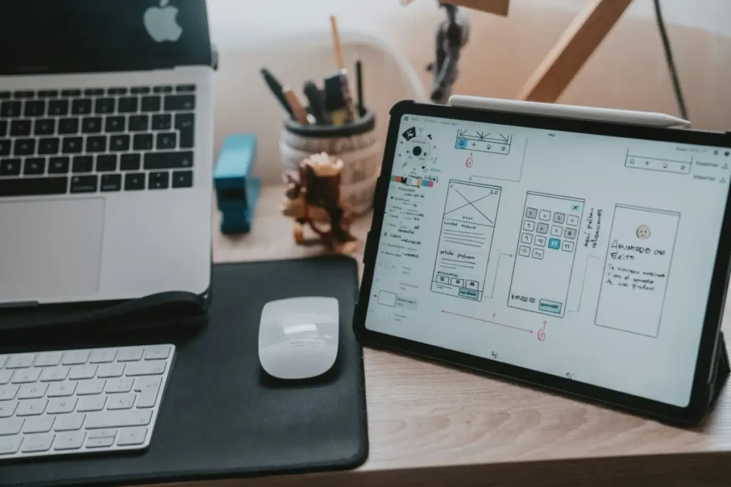 iPad の画面に UI ワイヤーフレームが表示されており、MacBook とマウスが並んだデザイナーの作業スペース