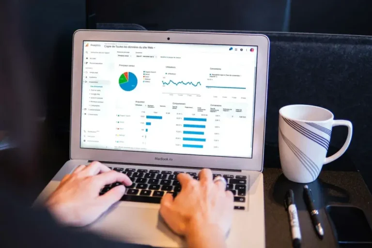 MacBook Air の画面に Google Analytics 風のダッシュボード、コーヒーカップと手元がキーボードに置かれている